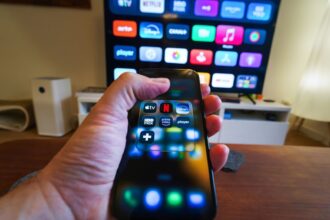 Close-up of a hand holding a phone displaying streaming apps in front of a TV with multiple app icons.
